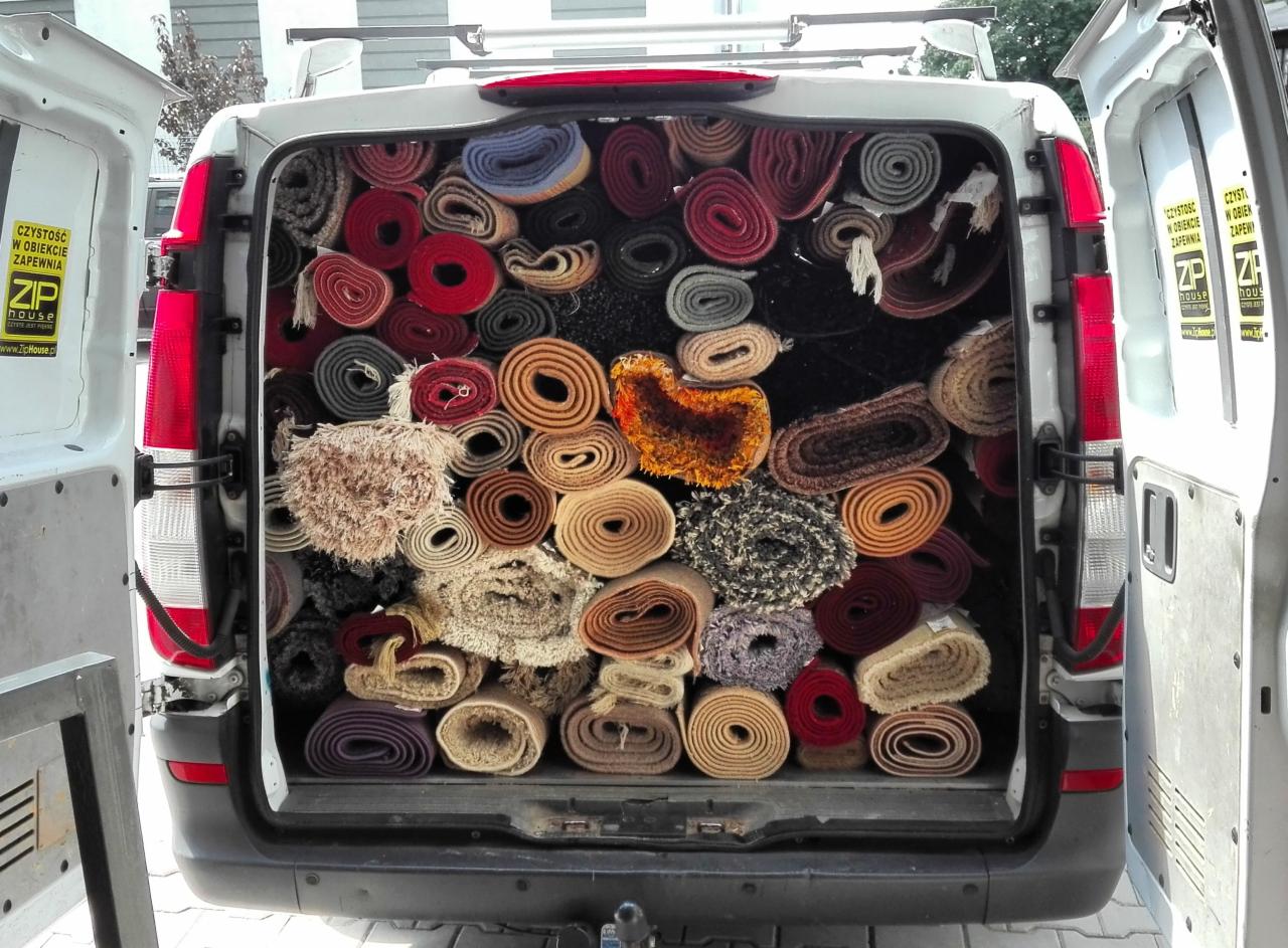 Wnętrze furgonetki wypełnionej zwiniętymi dywanami w różnych kolorach i wzorach, widok z tyłu pojazdu z otwartymi drzwiami, na drzwiach naklejki firmy sprzątającej.