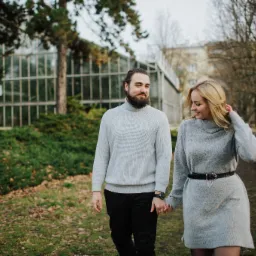 Para, mężczyzna z brodą w szarym swetrze i czarnych spodniach oraz blondynka w szarej sukience z paskiem, spacerują trzymając się za ręce w parku z budynkiem w tle.