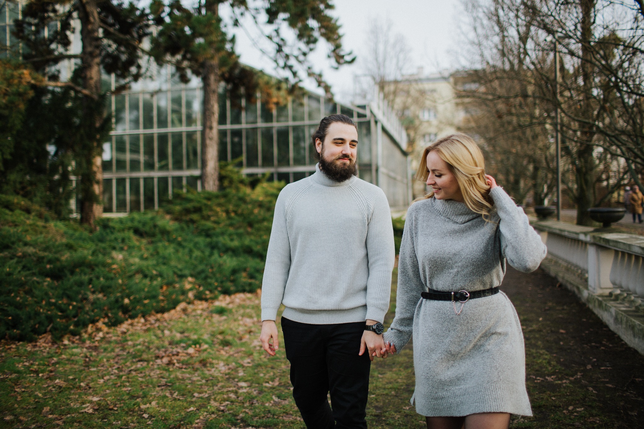 Para, mężczyzna z brodą w szarym swetrze i czarnych spodniach oraz blondynka w szarej sukience z paskiem, spacerują trzymając się za ręce w parku z budynkiem w tle.