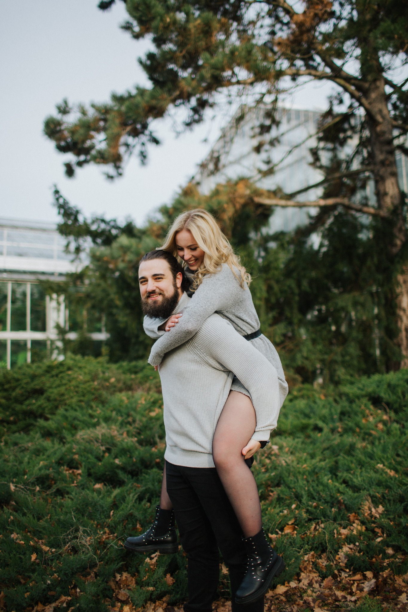 Uśmiechnięta blondynka na barana u brodatego mężczyzny w parku, w tle szklarnia i drzewa.