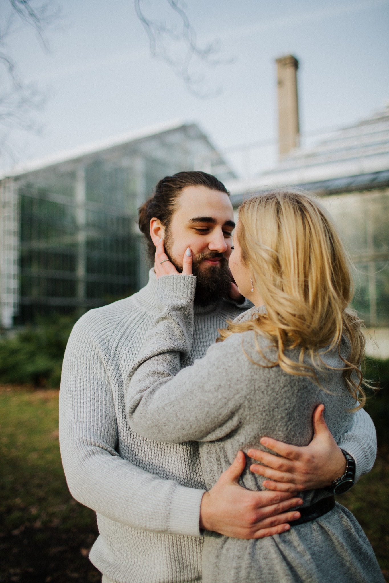 Para zakochanych w objęciach na tle szklarni, kobieta dotyka twarzy mężczyzny, oboje w szarych swetrach.