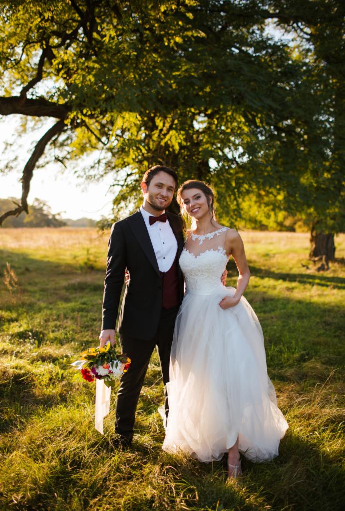 Para młoda w plenerze, on w smokingu z bordową muchą i kamizelką, ona w białej sukni z koronkową górą, trzyma bukiet, w tle łąka i rozłożyste drzewo w promieniach zachodzącego słońca.