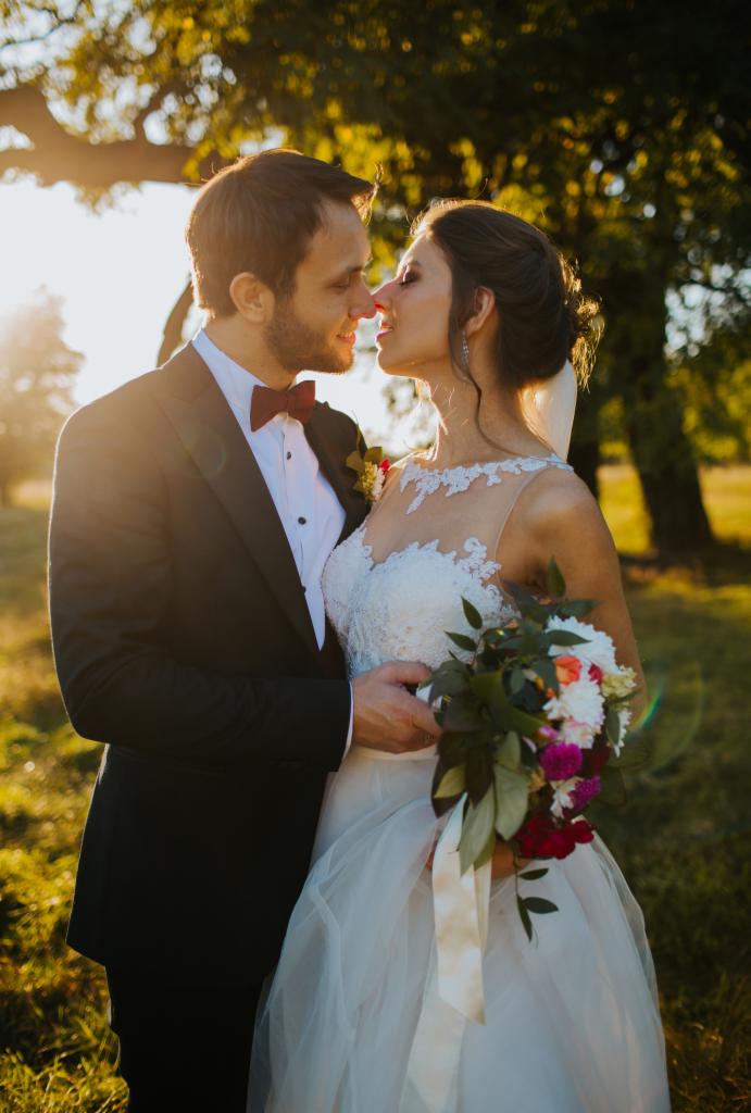 Para młoda, zbliżona twarzami w intymnym ujęciu na tle drzew i promieni słonecznych, panna młoda trzyma bukiet z białymi, różowymi i czerwonymi kwiatami, pan młody w czarnym smokingu z bordową muchą.