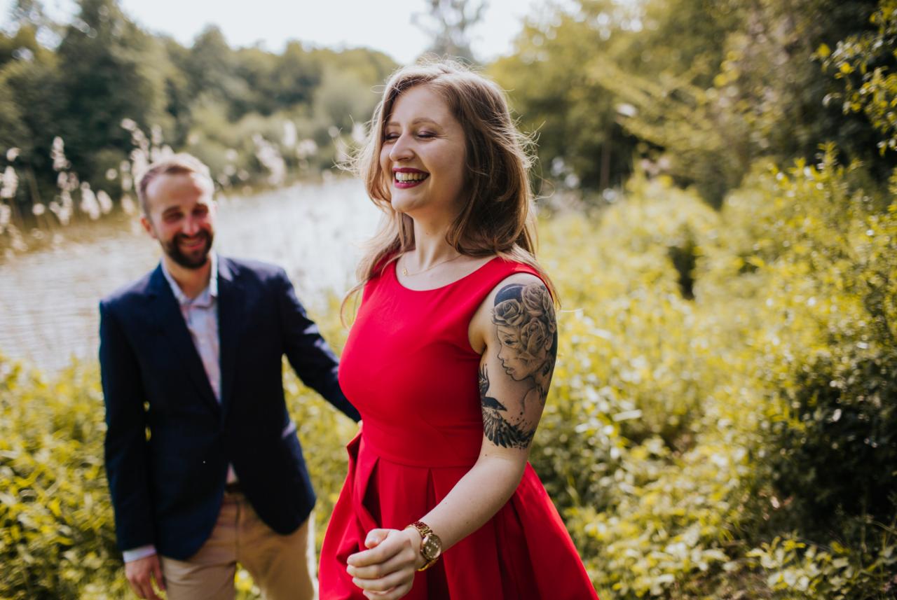 Uśmiechnięta kobieta w czerwonej sukience z tatuażem na ramieniu, spacerująca z mężczyzną w marynarce nad brzegiem jeziora.