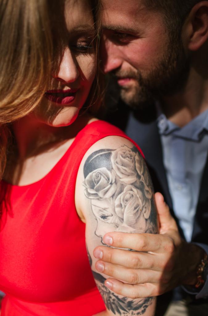 Para w zbliżeniu: Kobieta w czerwonej sukience z tatuażem róży na ramieniu, mężczyzna w niebieskiej koszuli i marynarce obejmuje ją, oboje z delikatnym uśmiechem.