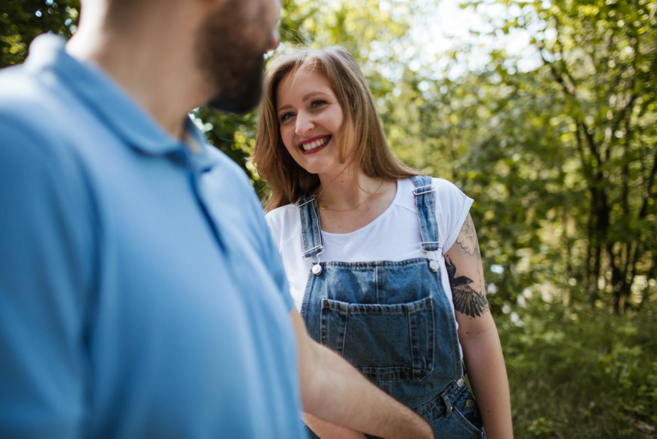 Uśmiechnięta kobieta w dżinsowych ogrodniczkach i białej koszulce, z tatuażem ptaka na ramieniu, patrzy na mężczyznę w niebieskiej koszulce polo na tle zieleni.