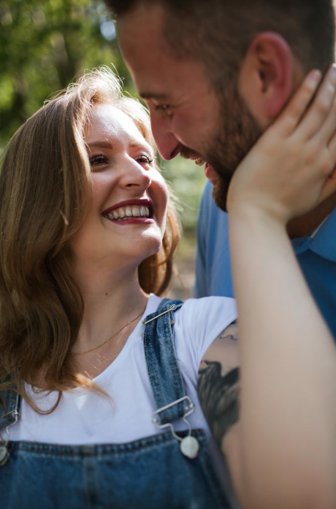 Uśmiechnięta kobieta w dżinsowych ogrodniczkach i białym t-shircie, z tatuażem na ramieniu, patrzy z czułością na mężczyznę z brodą, który trzyma jej twarz w dłoniach.