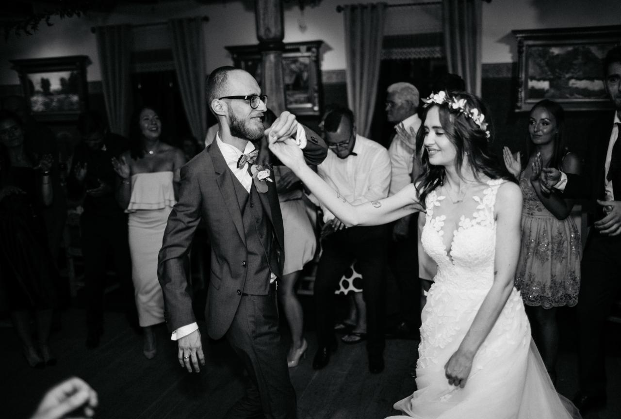 Czarno-białe zdjęcie pary młodej tańczącej na parkiecie podczas wesela, otoczonej przez gości klaszczących w dłonie. Panna młoda w koronkowej sukni i wianku, pan młody w garniturze z muszką. W tle...