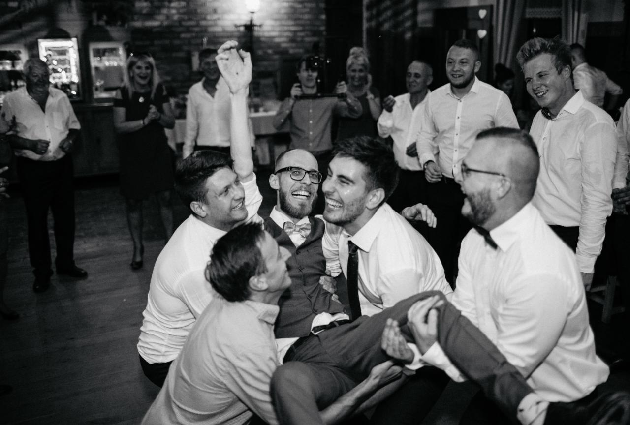 Czarno-białe zdjęcie uśmiechniętego pana młodego w okularach i muszce, podrzucanego przez grupę roześmianych mężczyzn na weselu, z gośćmi w tle.