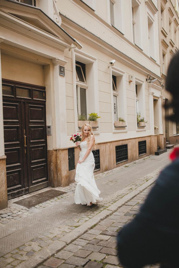 Panna młoda w białej sukni, trzymająca bukiet kwiatów, pozuje na brukowanej uliczce w Poznaniu, na tle kamienicy z jasną elewacją i ciemnymi, drewnianymi drzwiami.