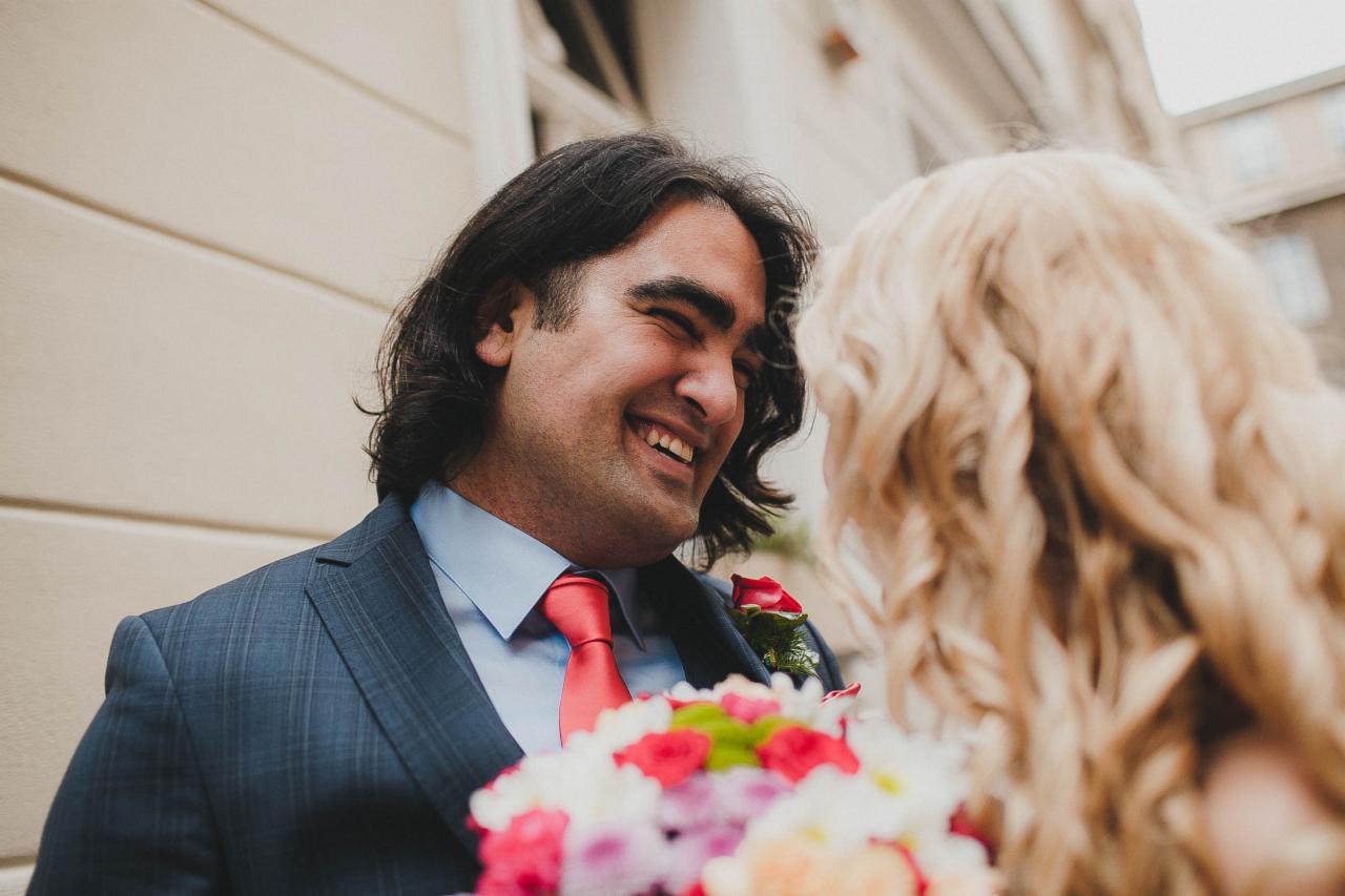 Uśmiechnięty pan młody w granatowym garniturze i czerwonym krawacie patrzy na pannę młodą z blond lokami, trzymając bukiet kolorowych kwiatów.