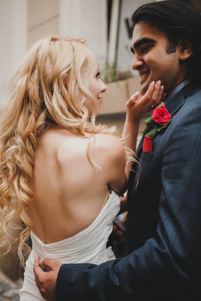 Panna młoda w białej sukni z odkrytymi plecami, spoglądająca z uśmiechem na pana młodego w granatowym garniturze z butonierką z czerwonej róży.