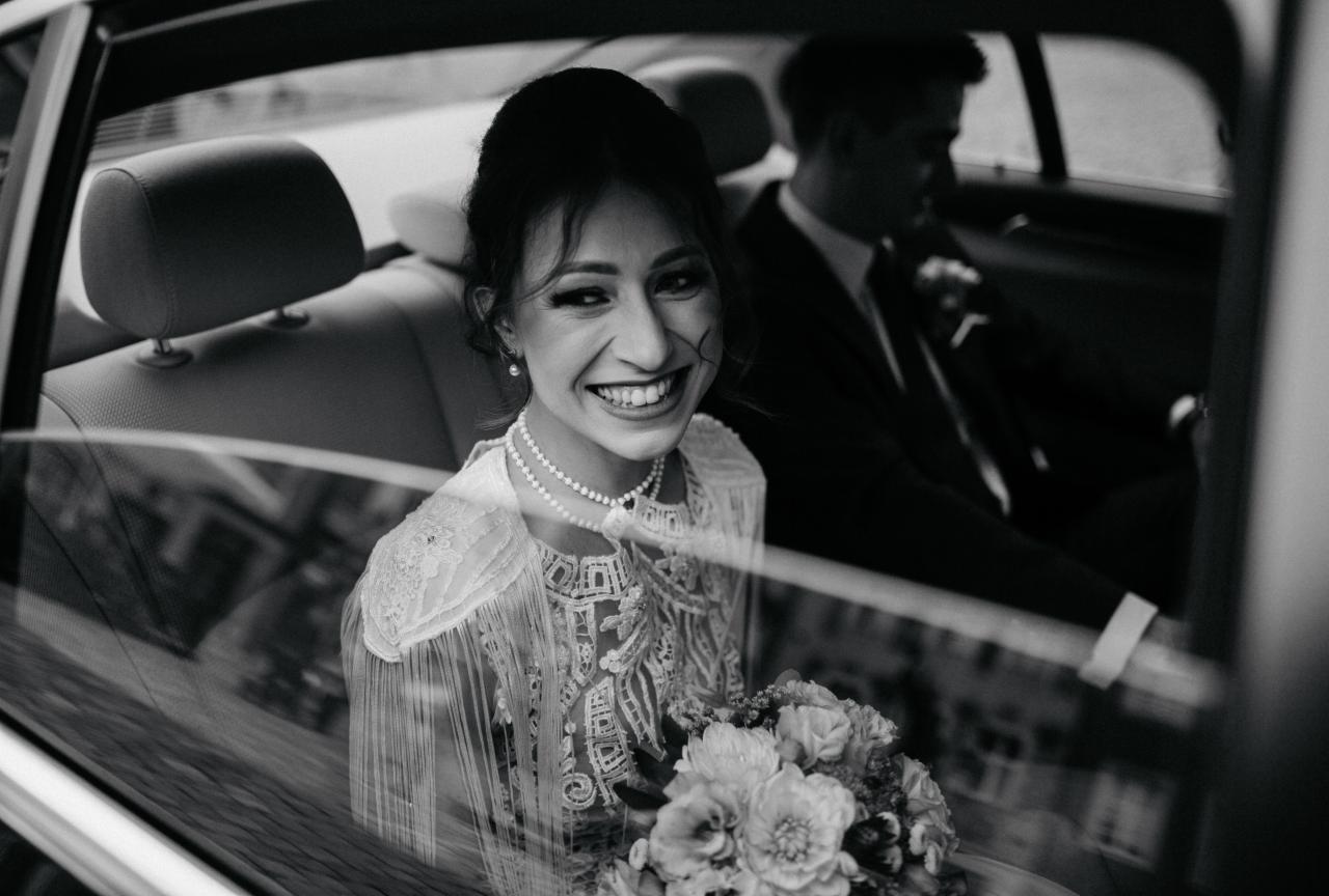 Czarno-białe zdjęcie uśmiechniętej panny młodej w samochodzie, z bukietem kwiatów, widoczny również pan młody w tle.