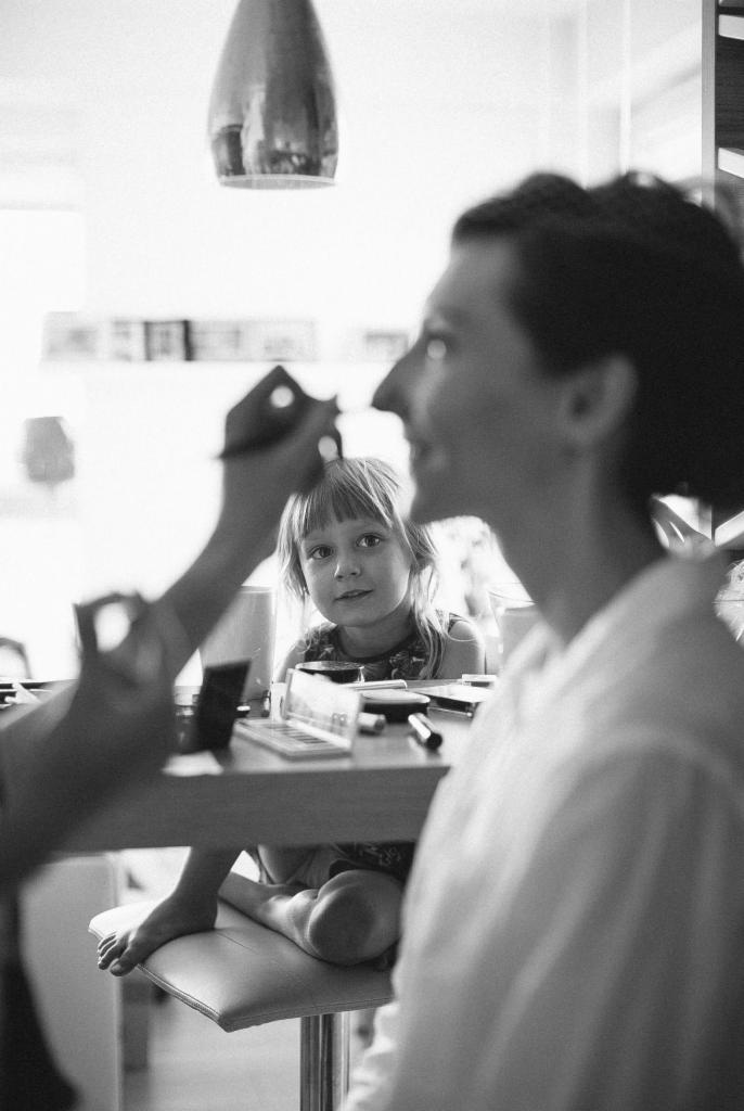 Czarno-białe zdjęcie kobiety, której makijaż wykonywany jest pędzlem, z zaciekawioną dziewczynką obserwującą proces w tle.