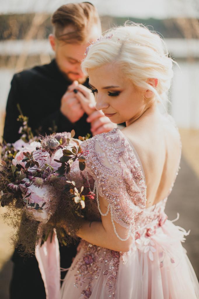Panna młoda w bogato zdobionej, pudrowo-różowej sukni z odkrytymi plecami, trzyma bukiet ślubny z ciemnymi akcentami, w tle narzeczony trzymający jej dłoń.
