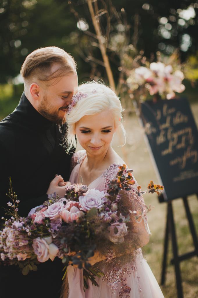Para młoda, blond kobieta w pudrowo-różowej sukni z bukietem fioletowych róż i mężczyzna w czarnym garniturze, stoją blisko siebie na tle tablicy z napisem.