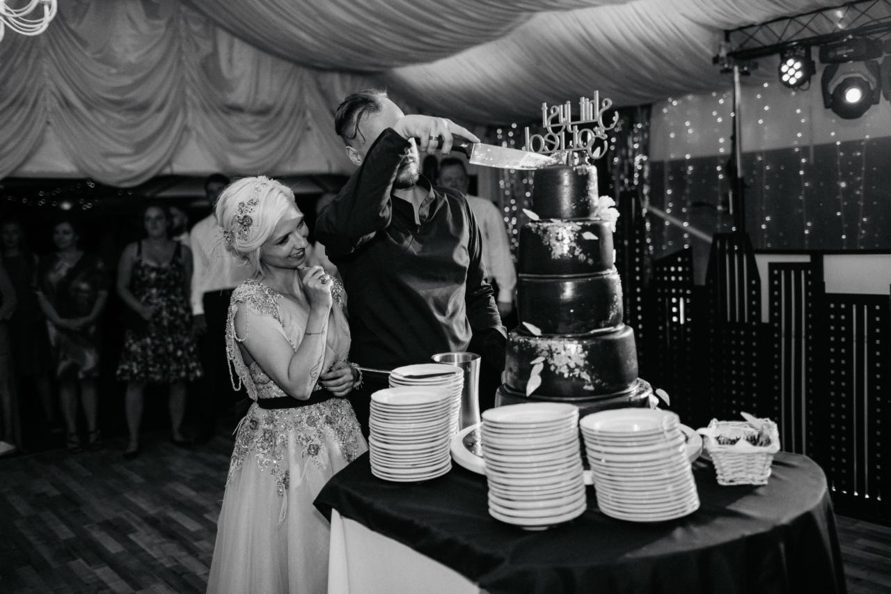 Czarno-białe zdjęcie pary młodej krojącej tort weselny; panna młoda uśmiecha się, obserwując; w tle dekoracje i goście.