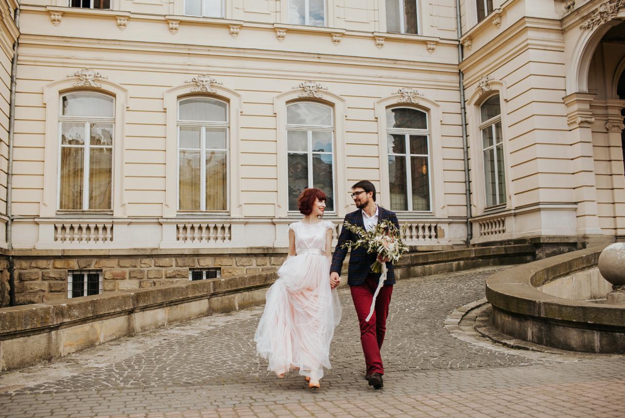 Para młoda spaceruje po brukowanej drodze przed klasycznym budynkiem z dużymi oknami; panna młoda w zwiewnej sukni z różowym odcieniem, pan młody w bordowych spodniach i marynarce w kratę trzyma...