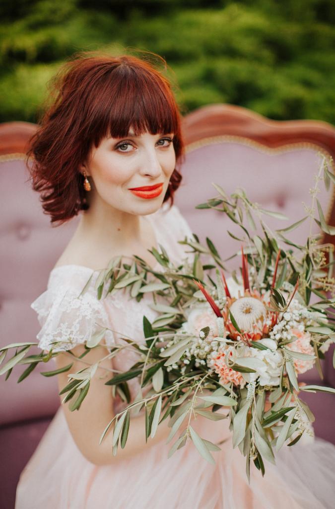Portret kobiety z rudymi włosami i czerwonymi ustami trzymającej bukiet kwiatów na tle różowej sofy i zieleni