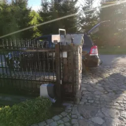 Automatyczna brama wjazdowa z czarnymi metalowymi przęsłami i kamiennymi słupkami, widoczny napęd bramy, zaparkowany samochód z otwartym bagażnikiem na brukowanej drodze, iglaki w tle, silne...