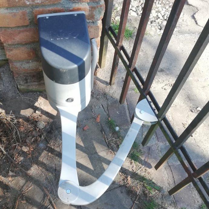 Szary automat do bramy marki NICE zamontowany na ceglanym słupku, połączony z metalową bramą wjazdową, widok z bliska na mechanizm.