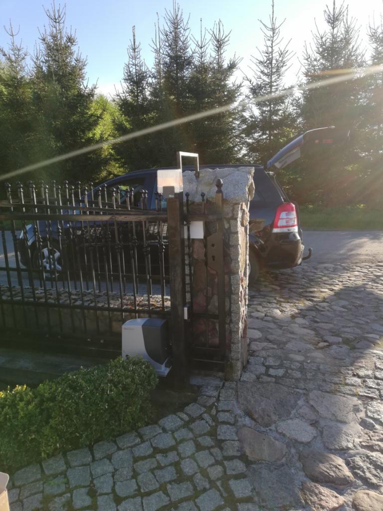 Automatyczna brama wjazdowa z czarnymi metalowymi przęsłami i kamiennymi słupkami, widoczny napęd bramy, zaparkowany samochód z otwartym bagażnikiem na brukowanej drodze, iglaki w tle, silne...