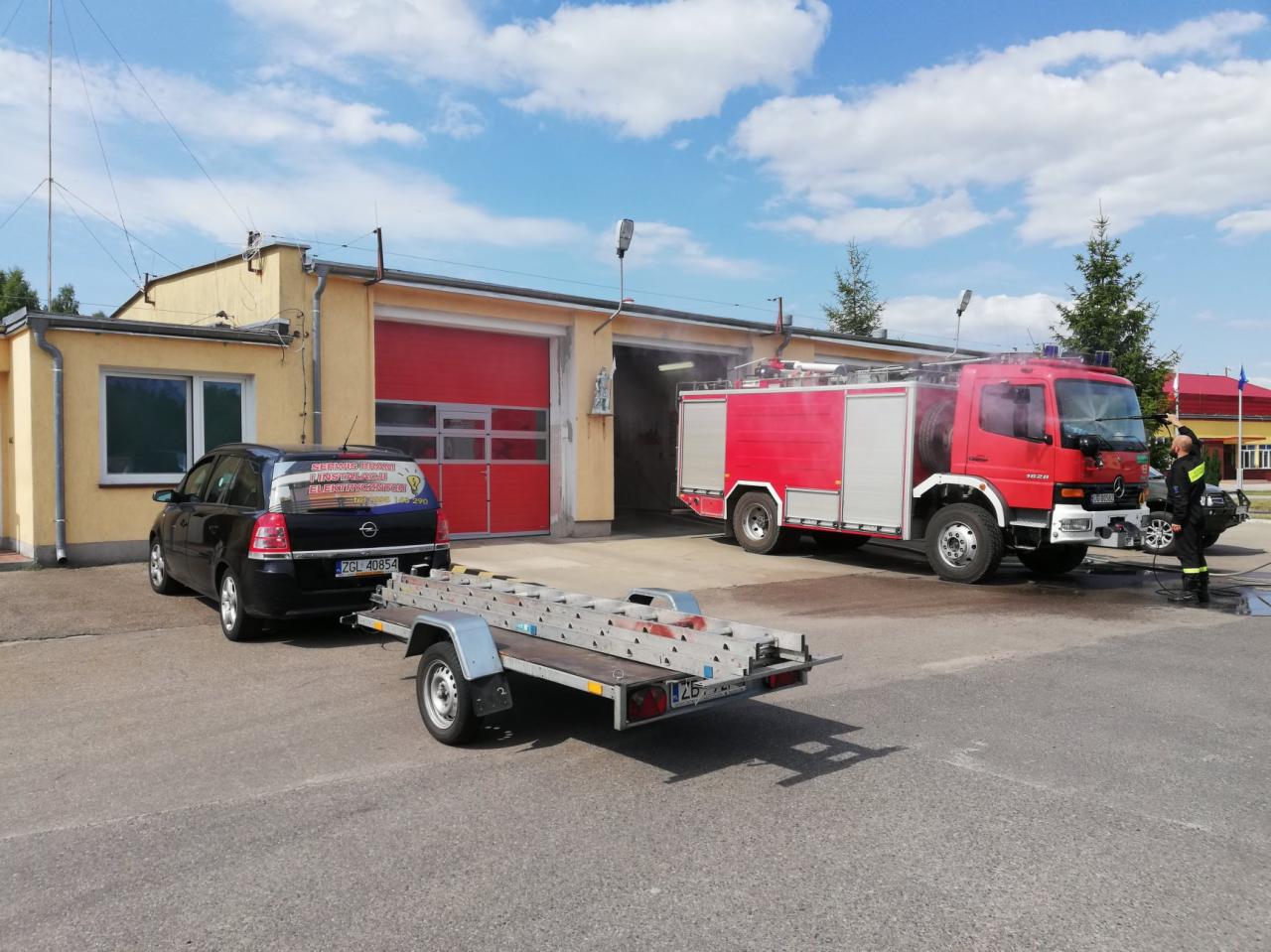 Czerwony wóz strażacki z otwartymi drzwiami garażowymi w tle, obok samochód osobowy z przyczepą zawierającą drabinę, strażak w umundurowaniu stoi obok wozu.