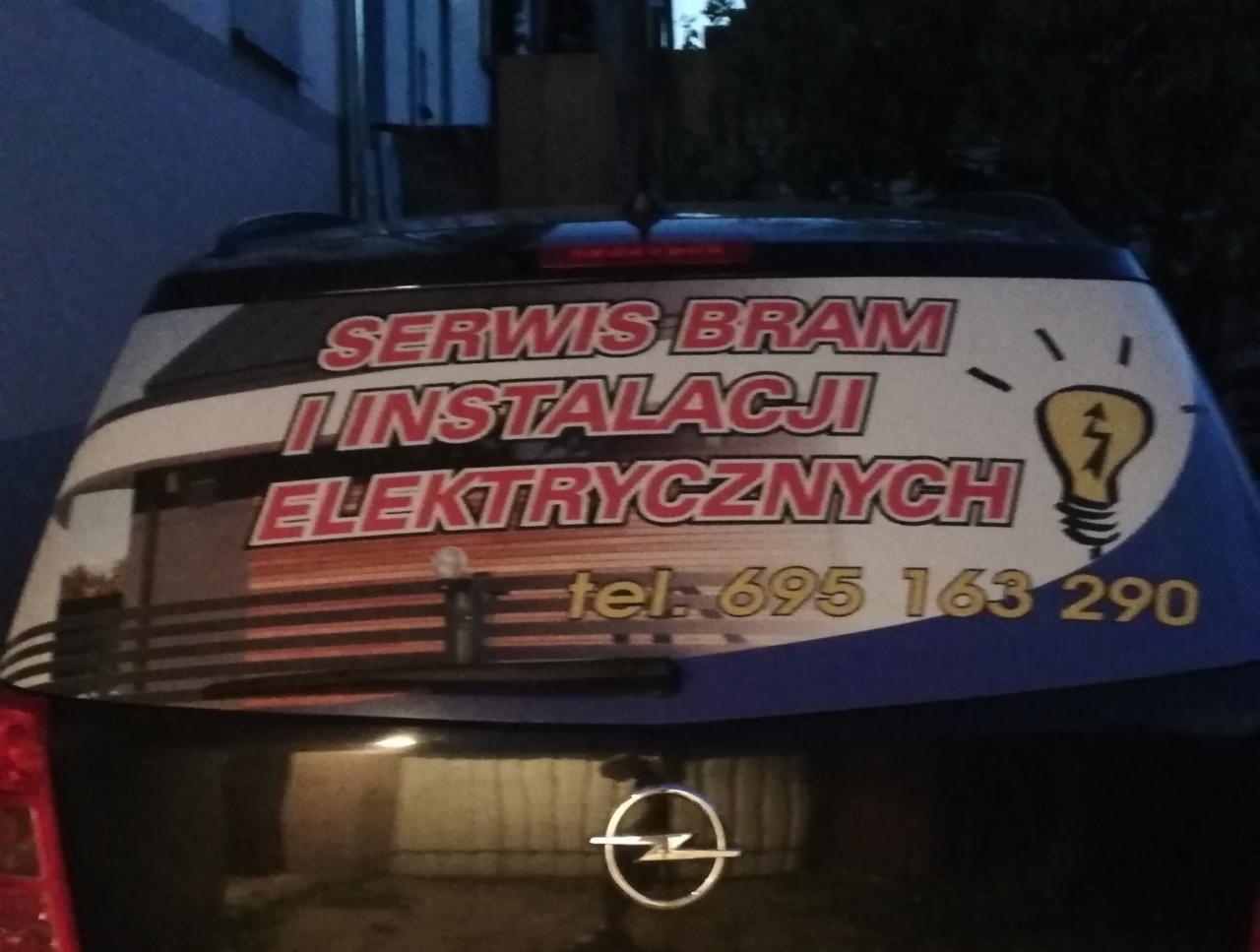 Reklama usług na tylnej szybie samochodu: Serwis Bram i Instalacji Elektrycznych, numer telefonu oraz grafika żarówki z błyskawicą.