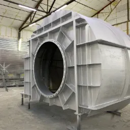 Duży, metalowy element konstrukcyjny z okrągłym otworem, świeżo pokryty srebrną powłoką, stojący w hali produkcyjnej; widoczne podpory i drabina w tle.