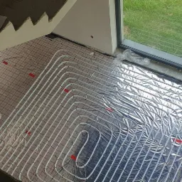 Montaż ogrzewania podłogowego: białe rurki ułożone na siatce, częściowo pokryte folią aluminiową, widok z góry, obok schody i okno z widokiem na trawnik.