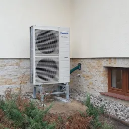 Zewnętrzna jednostka klimatyzacyjna Panasonic Inverter na metalowym stelażu przy ścianie budynku z dekoracyjną okładziną z kamienia piaskowego i zielenią w tle.