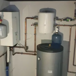 Zestaw urządzeń grzewczych w kotłowni: zasobnik ciepłej wody użytkowej z niebieską osłoną, bojler, ekspansomat i piec, połączone rurami miedzianymi.