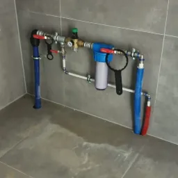 Instalacja wodna z filtrem mechanicznym, zaworami i rurami z tworzywa sztucznego, zamontowana na szarej, płytkowej ścianie w rogu pomieszczenia. Na podłodze widoczne ślady wilgoci.