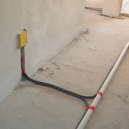 Surowy stan instalacji: Dwa czarne przewody wychodzące ze ściany, zabezpieczone czerwoną taśmą, biegną wzdłuż podłogi obok białej rury z czerwonymi oznaczeniami, w tle widoczne kolejne elementy...