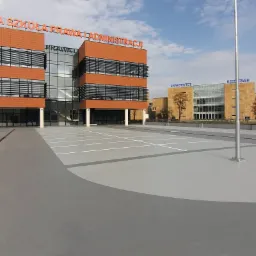 Szara, gładka nawierzchnia parkingu przed budynkami uczelni wyższych, z wyraźnie oznaczonymi białymi liniami stanowisk postojowych.