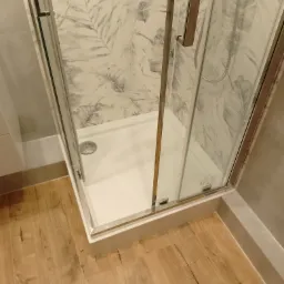 Kabina prysznicowa z brodzikiem i szklanymi drzwiami, wykończona płytkami z motywem liści, widoczna na tle drewnopodobnej podłogi w Tucholi.