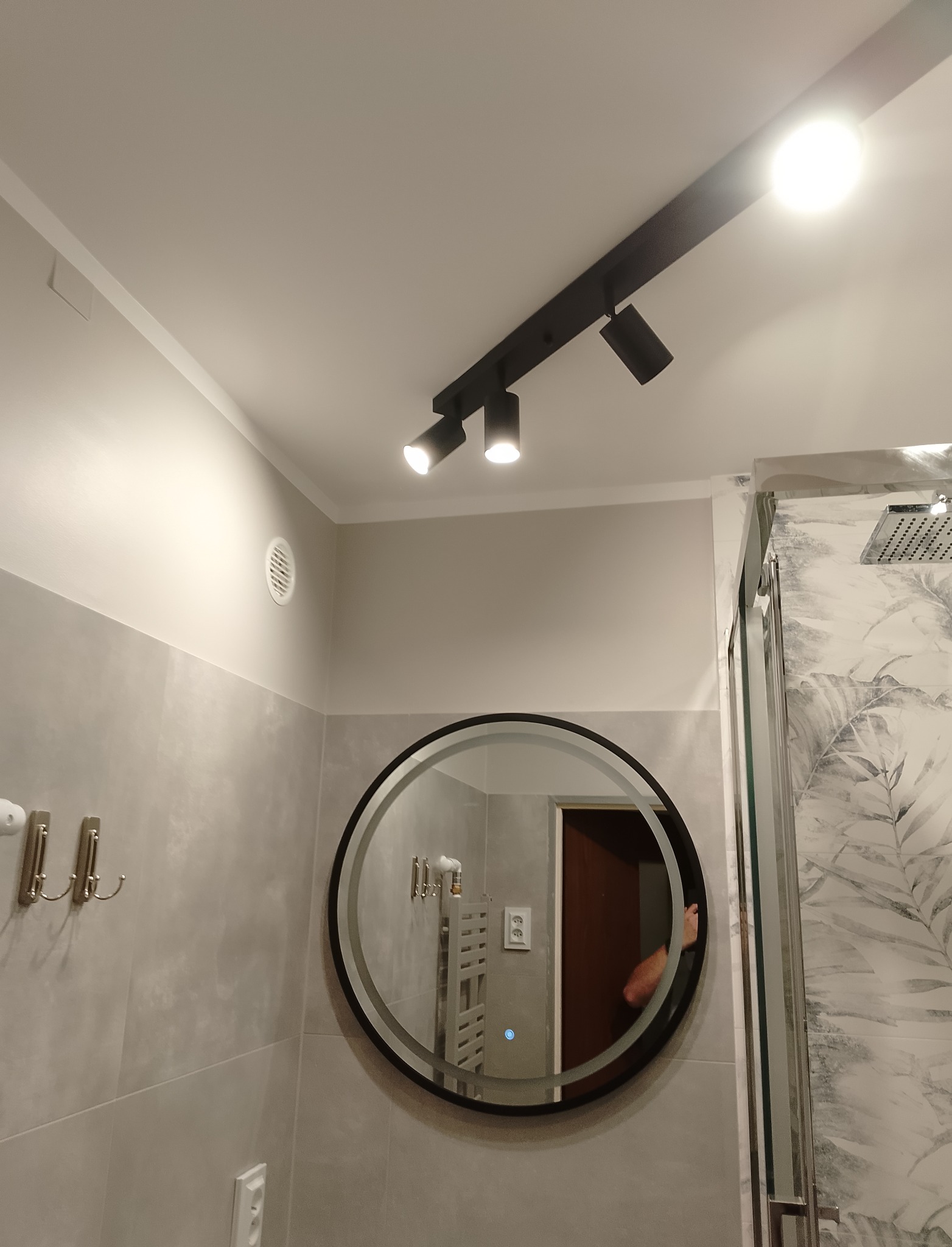 Nowoczesna łazienka z szarymi płytkami, okrągłym lustrem z podświetleniem LED, czarnym oświetleniem punktowym na suficie i kabiną prysznicową z marmurowym wzorem.