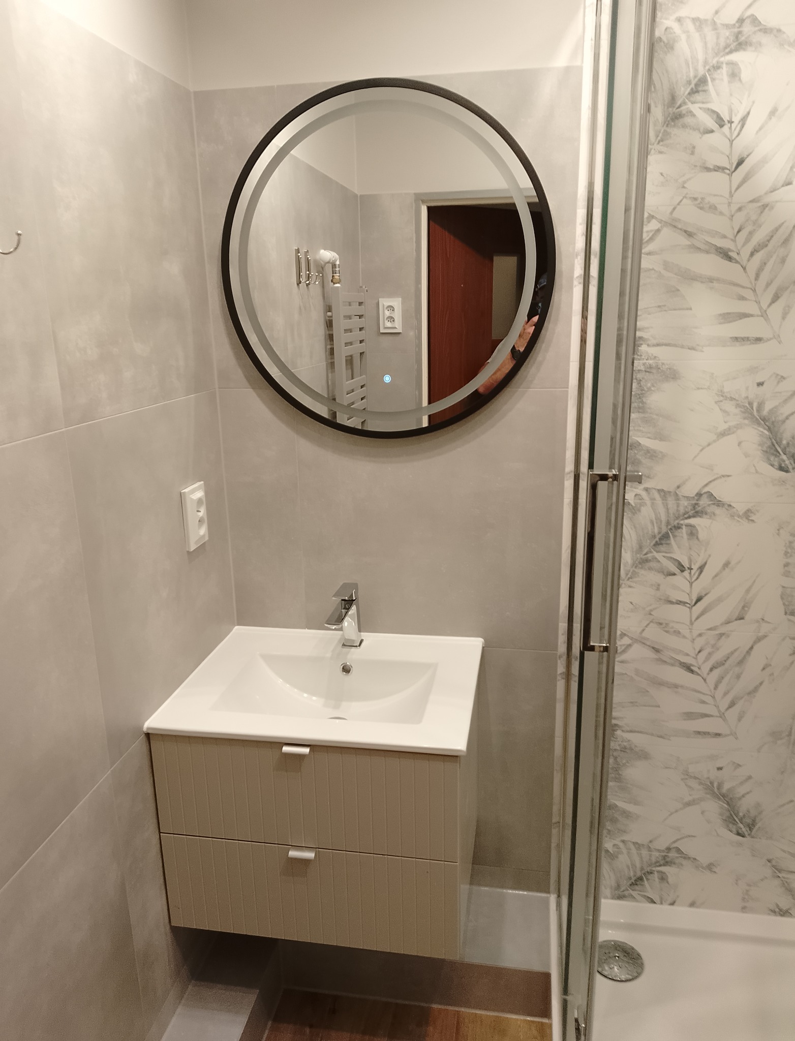 Nowoczesna łazienka z okrągłym lustrem podświetlanym LED, umywalką z szafką w odcieniach beżu i kabiną prysznicową z motywem liści na płytkach.