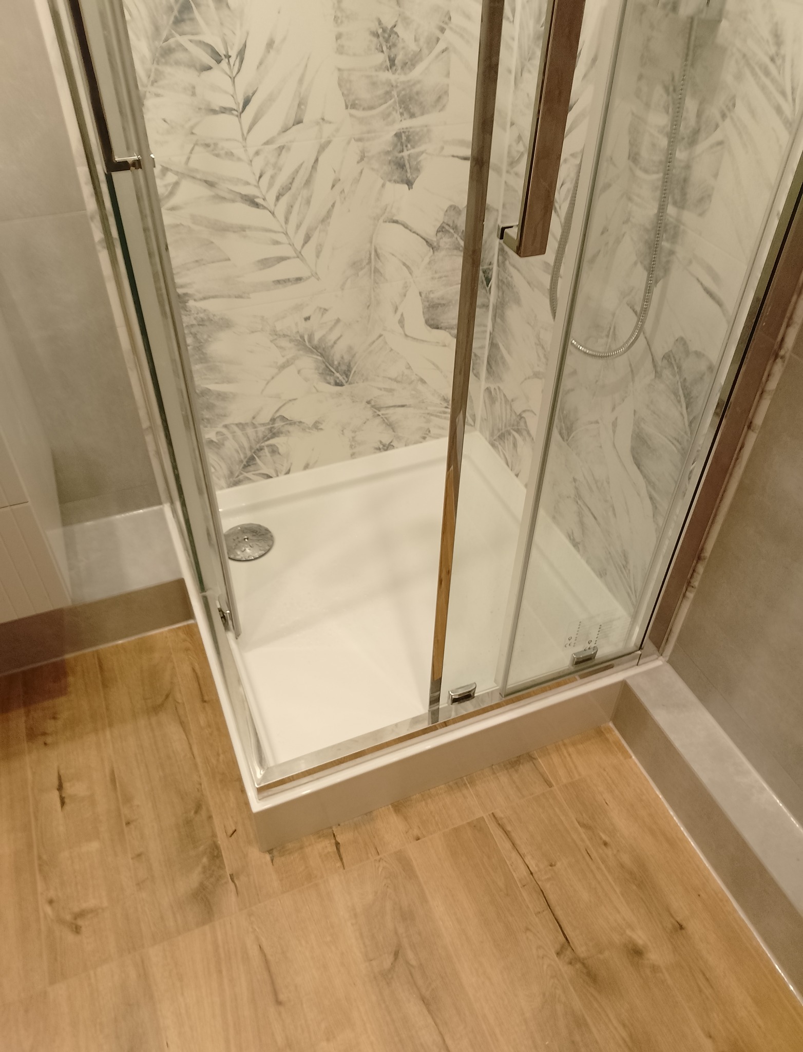 Kabina prysznicowa z brodzikiem i szklanymi drzwiami, wykończona płytkami z motywem liści, widoczna na tle drewnopodobnej podłogi w Tucholi.