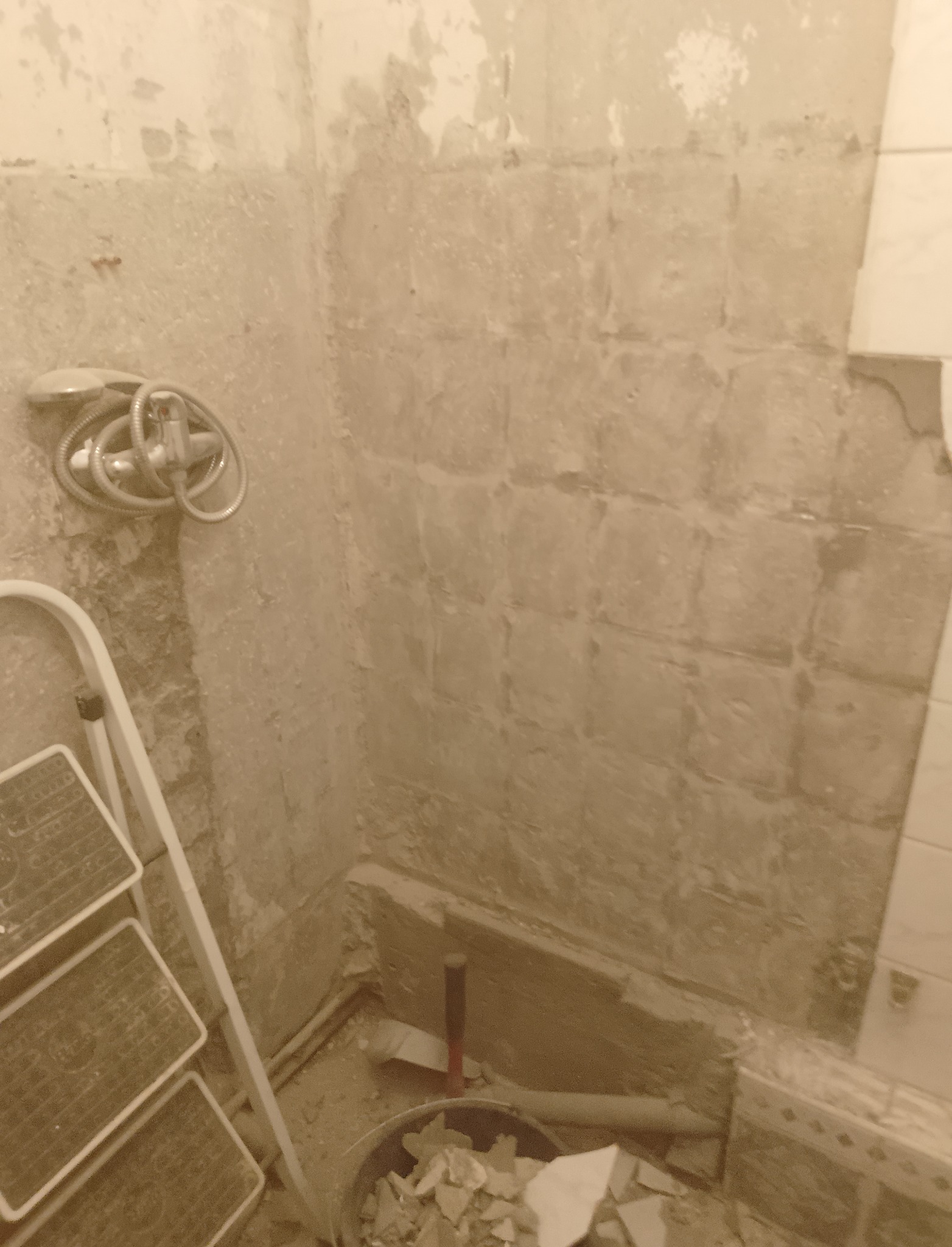 Demontaż płytek w łazience, widoczny fragment ściany z resztkami kleju, drabina oraz wiadro z gruzem i narzędzia.