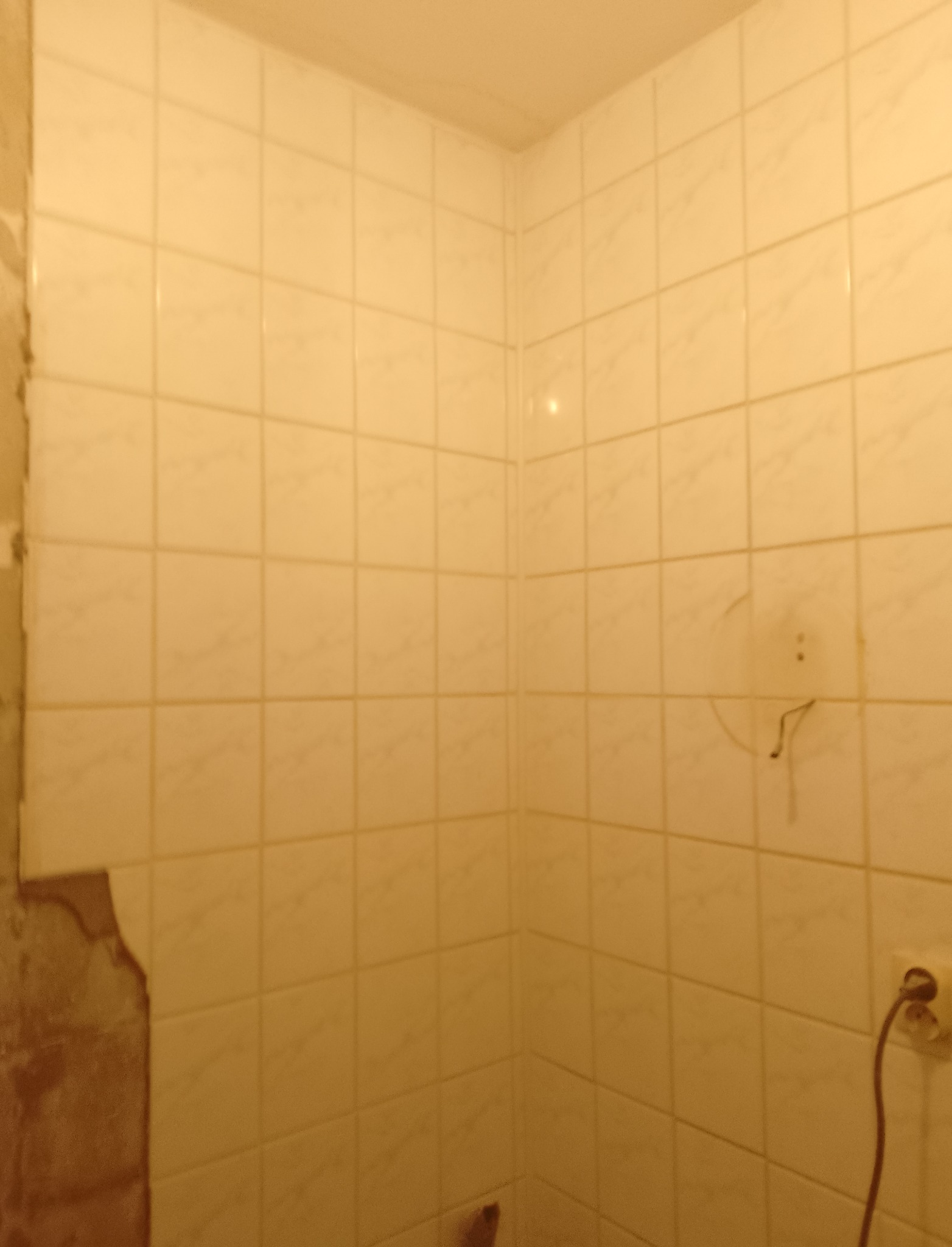 Ściana łazienki z częściowo usuniętymi, beżowymi płytkami, widoczne ślady po usuniętym mocowaniu i gniazdko elektryczne