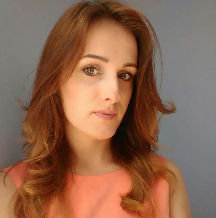 Portret kobiety z długimi, rudymi włosami, ubrana w koralową sukienkę, na jednolitym szarym tle.