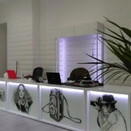 Biała lada z podświetleniem LED i grafikami kamer oraz portretu kobiety, laptop i akcesoria na blacie, w tle ściana z paneli 3D i roślina doniczkowa.