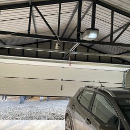 STALMAR MS - Otwarty garaż blaszany z bramą segmentową i zaparkowanym szarym samochodem na żwirowym podłożu, widoczny mechanizm otwierania bramy pod sufitem.