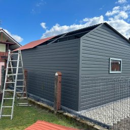 STALMAR MS - Montaż szarego budynku gospodarczego z elementami pokrycia dachu w kolorze ceglastym; widoczna drabina i pracownik na dachu, teren ogrodzony siatką.