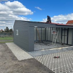 STALMAR MS - Montaż szarego garażu blaszanego na posesji z kostki brukowej; pracownik na dachu mocuje elementy pokrycia, widoczne drabiny i narzędzia.