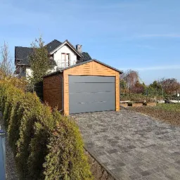 Nowoczesny, drewnopodobny garaż z szarą bramą segmentową, ustawiony na posesji z kostką brukową i żywopłotem, na tle domu z ciemnym dachem.