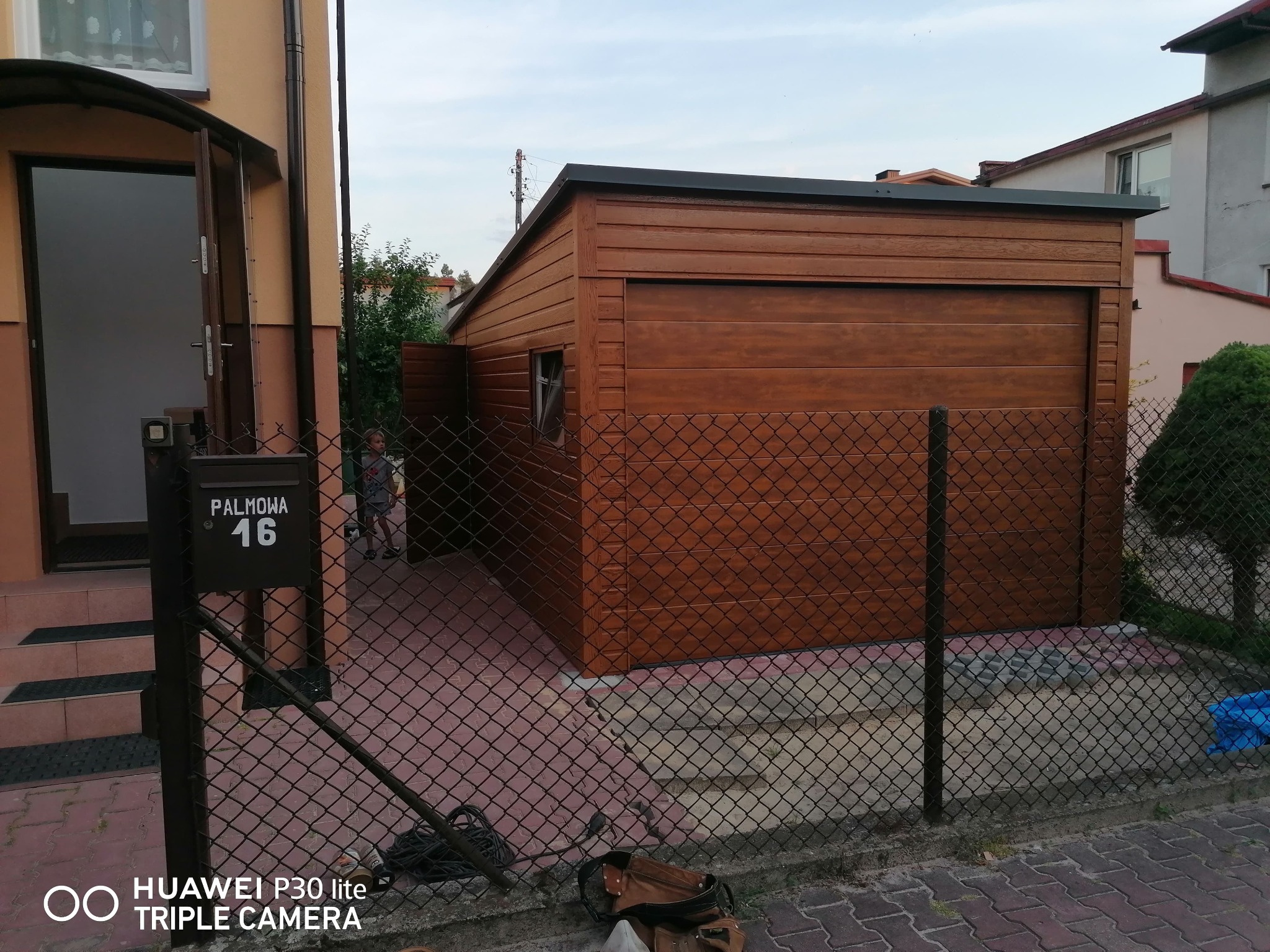 Nowy, drewnopodobny, brązowy budynek gospodarczy z płaskim dachem, widoczny przez siatkę ogrodzeniową na tle domu z otwartymi drzwiami wejściowymi i skrzynką pocztową o numerze 16.