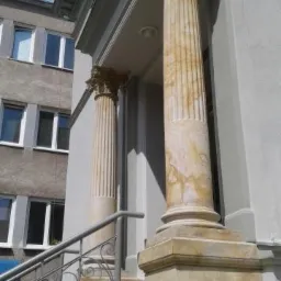 Odrestaurowane kolumny z kapitelami przed wejściem do budynku, widoczne ślady renowacji na kamieniu, metalowa balustrada schodów z ozdobnymi elementami.
