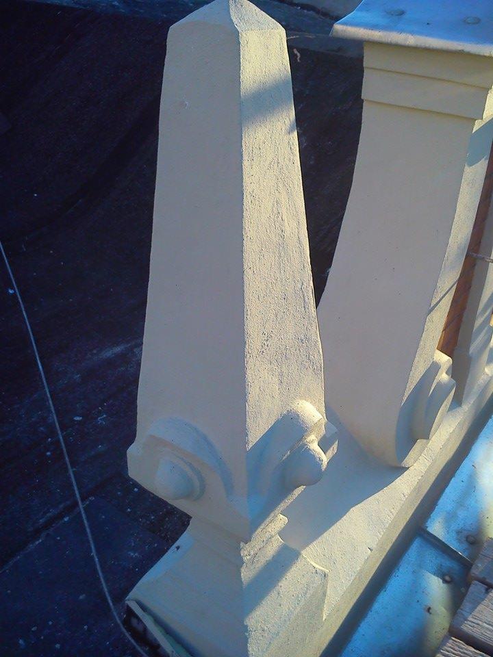 Odrestaurowany fragment balustrady z elementami dekoracyjnymi, widoczne ślady napraw i uzupełnień, oświetlony bocznym światłem słonecznym.